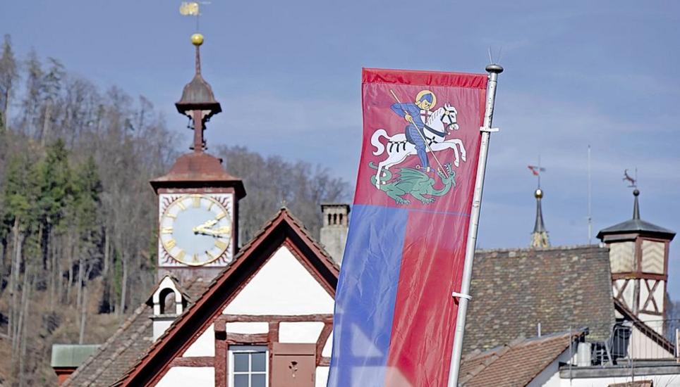In rot der blau gerüstete St. Georg mit gelbem Nimbus und Kreuzschild auf weissem Pferd, den grünen Drachen tötend.